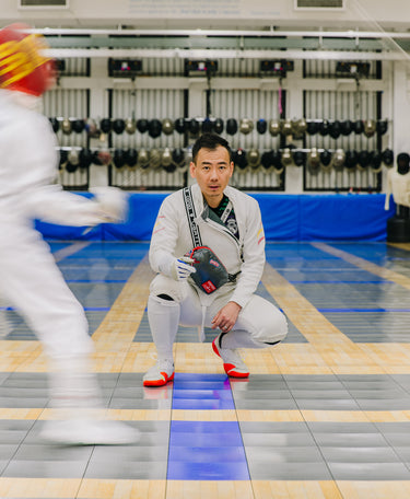 Fencer in a white outfit with red shoes and gloves, kneeling on a fencer's floor with blurred action in the foreground.