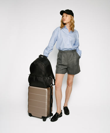 A woman stands against a light gray studio background wearing a light blue button-up shirt, gray shorts, black loafers, and a black cap, while holding a stack of two suitcases—a black backpack on top of a taupe hard-shell suitcase.