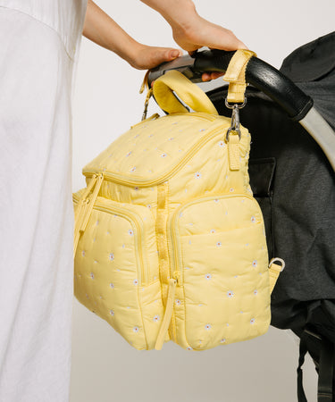 A pale yellow quilted diaper backpack with small embroidered floral details, held by a person next to a black stroller.