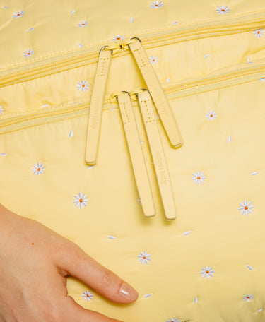 Close-up of a pale yellow quilted puffer bag with a daisy print, featuring dual yellow zippers with matching pull tabs and a hand pointing to the fabric texture.