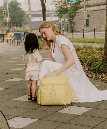 A woman in a white dress kneels on a city sidewalk next to a young child, holding a pale yellow quilted backpack with white polka dots. The scene is set in an urban outdoor environment with trees, street elements, and benches in the background.
