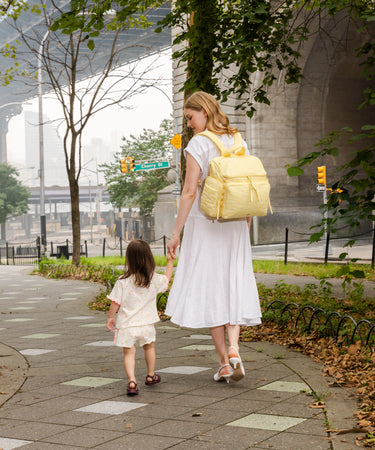 A woman in a white dress and a young girl in a matching cream outfit walking hand-in-hand on a city sidewalk, with the woman wearing a pale yellow backpack. Trees, a stone bridge, and urban background are visible.