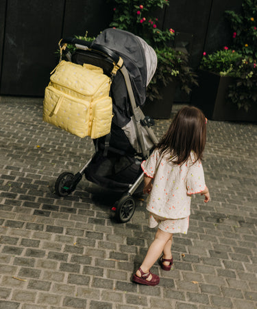 A toddler in a white floral romper walks beside a black stroller with a large yellow padded bag attached to the handle, on a gray brick pathway with potted plants in the background.