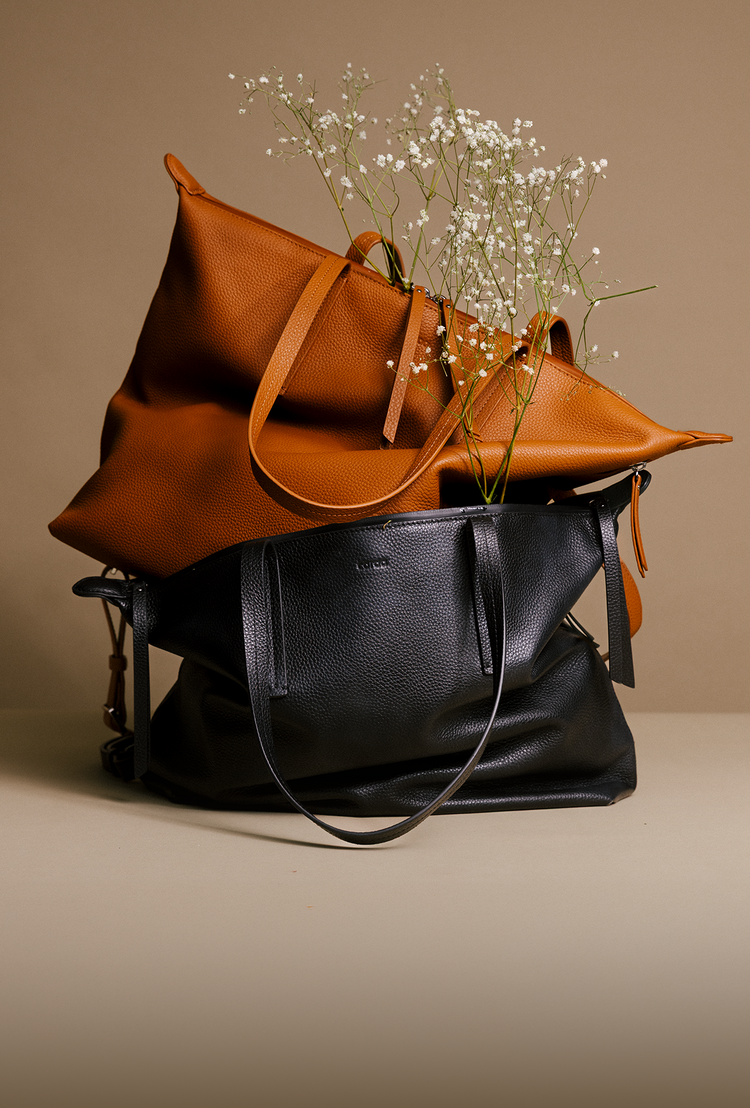 Two leather tote bags stacked on a beige surface against a warm beige background, with small white baby's breath flowers placed on top of the upper bag. The top bag is a warm camel/cognac brown, and the bottom bag is black.