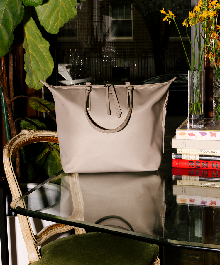 A large structured tote bag in light beige with metallic silver handles sits on a glass table in a styled interior setting with a chair, books, and flowers.