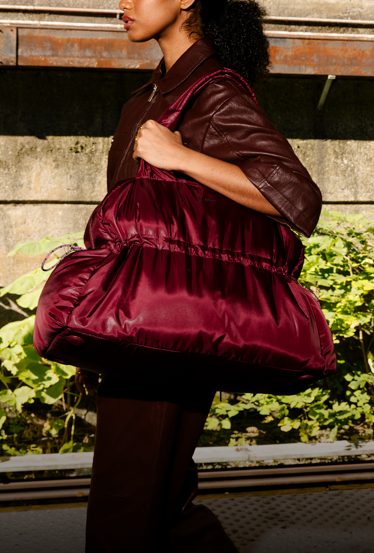 A person in a matching dark burgundy jumpsuit holds a large shiny burgundy duffel bag resting on top of a silver suitcase, standing on a train platform beside tracks at sunset.