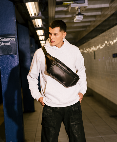 Man wearing a white hoodie and black pants with a black crossbody bag in a subway station.