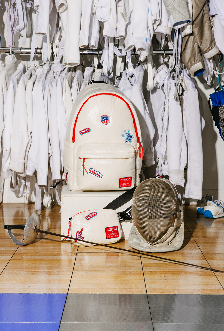 White backpack with colorful patches on a tiled floor in a room with hanging clothes and sports equipment.