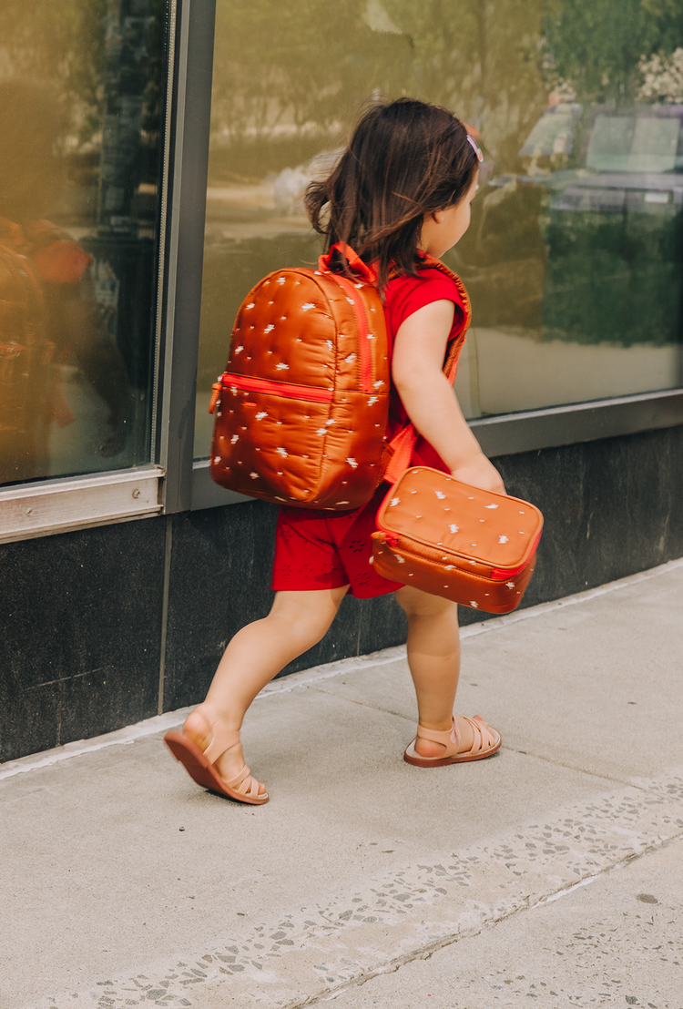 A child in a red matching two-piece outfit holds a small red bag while standing next to an adult wearing olive green pants and holding a large red duffel bag with gold star details, set against a modern building facade.