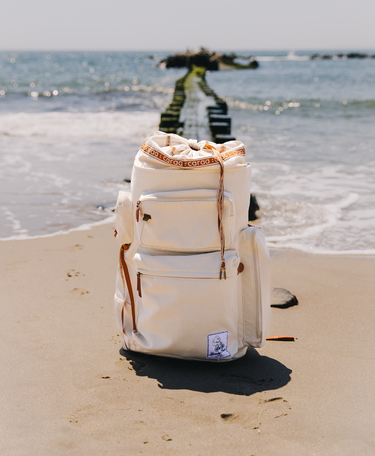 Beige backpack with brown straps on a sandy beach with ocean in the background