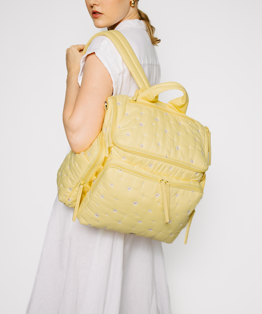 A woman wearing a white sleeveless dress carries a pale yellow quilted backpack with multiple zip compartments and top handles against a plain light gray studio background.