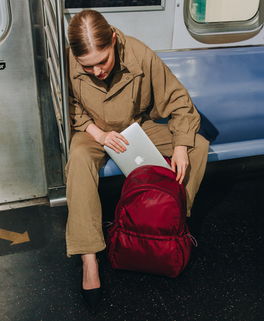 A woman in a brown jumpsuit sits on a subway train next to a blue bench, holding a silver laptop next to a deep red backpack.
