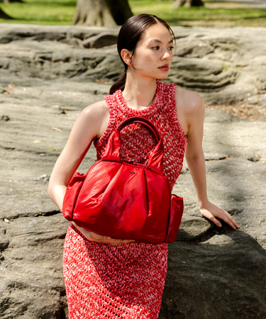 A woman outdoors wearing a red-and-white textured dress and carrying a large shiny red handbag, posed against large weathered rocks.