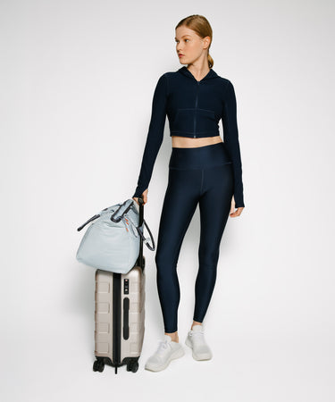 A woman stands in a studio wearing a dark navy athletic matching set consisting of a cropped zip-up hoodie and high-waisted leggings, holding a light blue duffel bag on top of a silver suitcase against a plain light gray background.