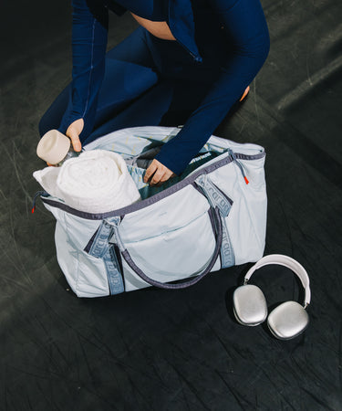 A person in a navy blue activewear set packs items into a large light blue-gray duffel bag with gray trim. A pair of white over-ear headphones rests on the dark floor beside the bag.