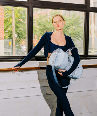 A woman in a navy blue activewear set stands by a window, holding a light blue-gray shoulder bag with a blue strap.