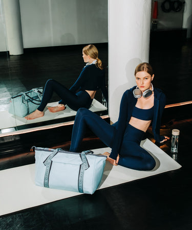 A woman in a navy blue activewear set sits on a white yoga mat in a gym, wearing silver headphones around her neck and a light blue gym bag beside her. Her reflection is visible in a large mirror behind her.