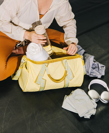 A pale yellow gym duffel bag with light blue patterned handles sits on a dark floor while a person packs it with a towel and water bottle. Folded clothing, white headphones, and a gray garment are placed nearby.