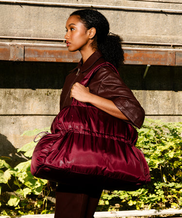 A woman in a dark burgundy leather jacket and matching pants carries a large, puffy satin-like shoulder bag in a deep burgundy shade, set against a concrete wall with green foliage.